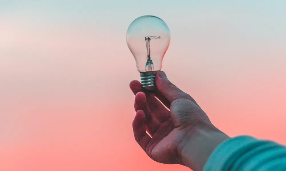 A lightbulb being held by someone wearing a blue sweater in front of a sky transitioning from blue to coral, Photo by Diego PH from Unsplash