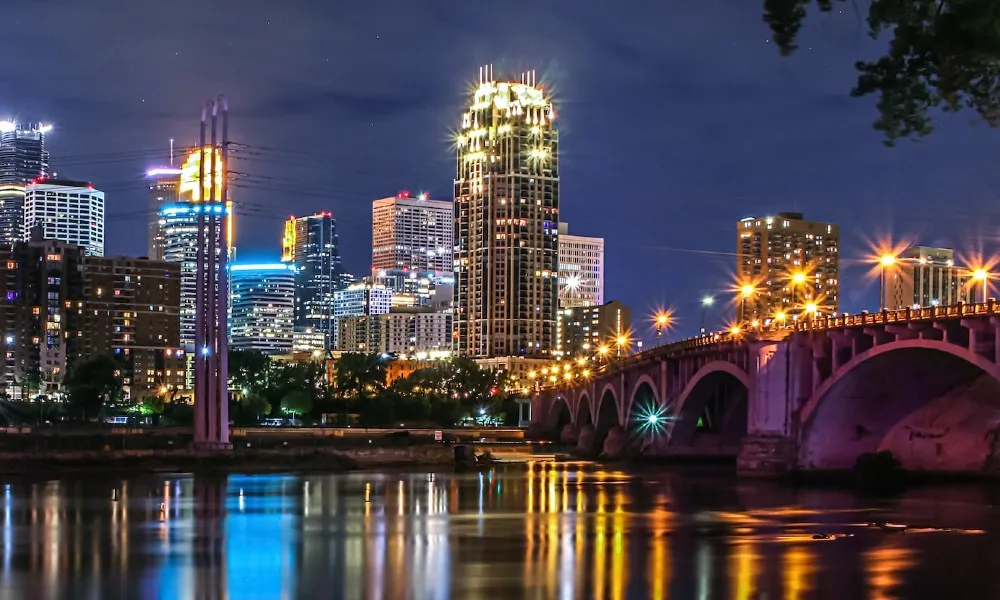 Minneapolis cityscape at night, bridge over river, lens flare from street and building lights.